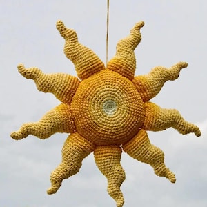 Könnte beinhalten: Ein gehäkeltes Sonnenornament mit einer runden, strukturierten Mitte in Gelb- und Orangetönen und einem weißen Kern. Die Sonne hat zehn gewellte, spitze Strahlen, die nach außen ragen, gefertigt in einem gelben Farbverlauf, aufgehängt an einer Schnur.