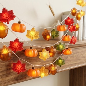 May include: A string of decorative LED lights featuring translucent plastic shapes of orange pumpkins, brown acorns, and maple leaves in red, yellow, and green. The lights are illuminated, creating a warm glow along the white wire strand.