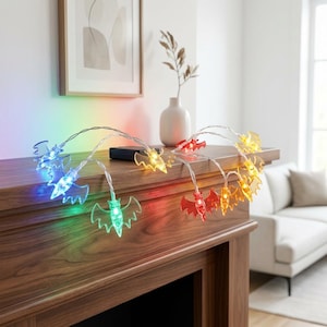 May include: A string of LED lights featuring translucent bat-shaped covers in a gradient of colors including blue, green, yellow, orange, and red. The lights are connected by a thin wire and powered by a small black rectangular battery pack.