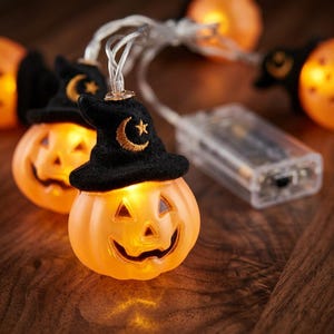 Puede incluir: Una cadena de luces LED con calabazas de plástico naranja que llevan sombreros de bruja de fieltro negro. Cada sombrero está bordado con una luna creciente y una estrella doradas. Las luces están conectadas por un cable transparente.