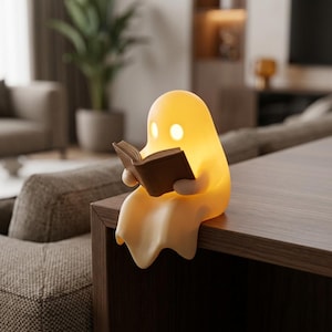 May include: A soft, glowing, pale yellow ghost-shaped lamp sitting on a flat surface. The ghost figure has two bright, circular illuminated eyes and is holding a small, brown, open book in its hands, appearing to read the pages.