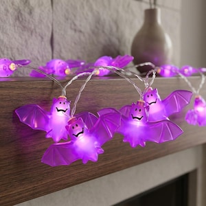 May include: A string of LED lights featuring translucent purple bat-shaped covers. Each bat has a smiling face and is illuminated from within by a bright light. The bats are connected by a thin, clear wire, creating a decorative lighting display.