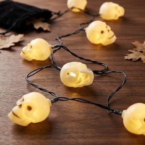 May include: A string of decorative lights featuring multiple off-white, hollow skull-shaped covers. Each skull contains a small, warm-toned LED bulb that emits a soft glow. The skulls are connected by a thin black wire, creating a continuous strand.
