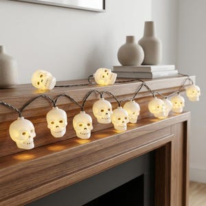May include: A string of white skull-shaped LED lights connected by a dark wire. Each individual skull features cutouts for the eyes and nose, emitting a warm, soft glow from within. The light strand is draped across a wooden surface.