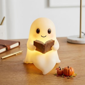 May include: A glowing, off-white, ghost-shaped lamp with a smooth, rounded head and a friendly face, holding a small, brown, open book. The lamp emits a soft, warm light, and the ghost features dark, oval eyes and a simple, curved smile.