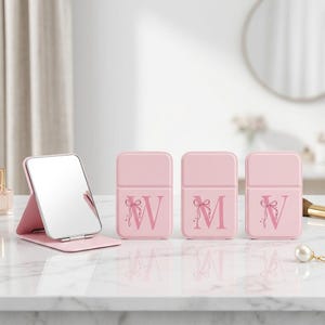 Puede incluir: Cuatro espejos plegables rectangulares de color rosa claro. Un espejo está abierto, mostrando la superficie reflectante. Los otros tres están cerrados, cada uno con una letra W, M o V en rosa oscuro y un pequeño lazo dibujado.