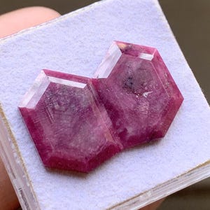 Könnte beinhalten: Zwei facettierte, tief pink-rote Rubine in sechseckiger Form. Die Edelsteine haben eine flache Oberseite mit abgeschrägten Kanten und weisen natürliche interne Einschlüsse sowie eine halb-undurchsichtige Textur im Kristallgefüge auf.