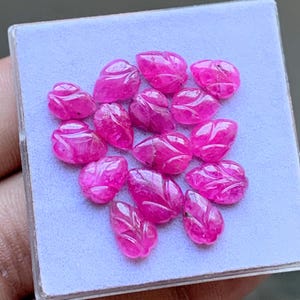 Könnte beinhalten: Eine Ansammlung von dreizehn kleinen, leuchtend pinkfarbenen Edelsteinen, die in Blattform geschnitzt sind. Jeder einzelne Stein weist detaillierte geätzte Adern und eine polierte Oberfläche auf und ist in einer Gruppe auf einem hellblauen quadratischen Display angeordnet.