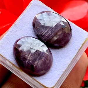 Puede incluir: Dos rubíes ovalados y facetados de tono rojo púrpura intenso con estrías internas visibles. Las piedras preciosas están colocadas una al lado de la otra sobre un inserto de espuma blanca texturizada dentro de una caja de plástico transparente.
