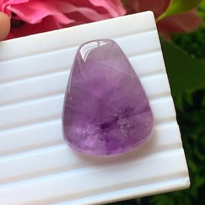 Könnte beinhalten: Ein polierter, tropfenförmiger Amethyst-Edelstein mit einem durchscheinenden violetten Farbton, sichtbaren inneren kristallinen Einschlüssen und einer glatten, abgerundeten Oberfläche auf einem weißen, gerillten Display.
