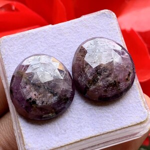 Pode incluir: Duas pedras preciosas redondas e facetadas com um tom púrpura e magenta profundo. As pedras apresentam uma superfície superior lapidada em rosa com inclusões internas visíveis e manchas minerais escuras no material translúcido e polido.