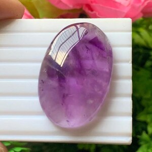 Könnte beinhalten: Ein ovaler Amethyst-Edelstein mit einer glatten, polierten Oberfläche. Der Stein zeigt eine durchscheinende violette Farbe mit natürlichen Variationen und Einschlüssen. Eine helle Lichtreflexion ist auf dem oberen Teil des Steins sichtbar.