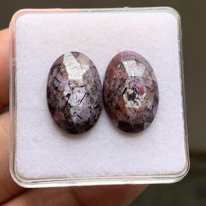 Könnte beinhalten: Zwei ovale, facettierte Edelsteine mit gesprenkeltem Aussehen in Violett-, Rosa- und Schwarztönen. Die Steine haben eine polierte Oberfläche mit sichtbaren Einschlüssen und dunklen, nadelartigen Mustern, präsentiert in einer quadratischen weißen Box.