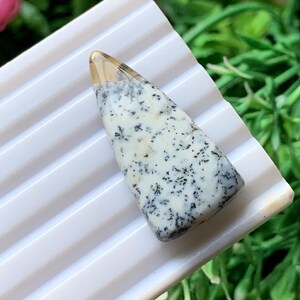 Könnte beinhalten: Ein dreieckiger Cabochon-Stein mit einer glatten, undurchsichtigen weißen Basis, die komplizierte, dunkelgraue dendritische farnartige Einschlüsse aufweist, sowie einer durchscheinenden, bernsteinfarbenen Spitze, die in den gemusterten weißen Körper übergeht.
