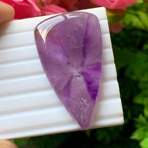 Könnte beinhalten: Ein polierter, tropfenförmiger Amethyst-Edelstein mit einem durchscheinenden violetten Farbton, natürlichen weißen Bändern und kristallinen Einschlüssen. Der Stein hat eine glatte, abgerundete Oberfläche und liegt auf einer weißen gerippten Basis.