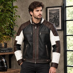 Puede incluir: Una chaqueta de cuero con bloques de color que presenta un panel central negro, hombros y ribetes de color marrón oscuro, y mangas blancas con detalles en negro. La chaqueta incluye un cuello alto con cierre de broche, cremallera y cuatro bolsillos.