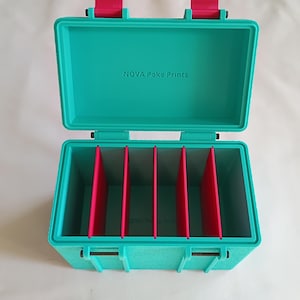 May include: A teal rectangular storage box with a hinged lid and bright pink latches. The interior features four vertical pink dividers creating five compartments. The text NOVA Poke Prints is printed in dark lettering on the inside lid.