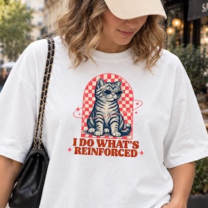 Könnte beinhalten: Ein weißes Kurzarm-T-Shirt mit einer Grafik einer Katze mit Sonnenbrille in einem rot-weiß karierten Bogen. Der Text unter der Katze lautet I DO WHAT'S REINFORCED. Das T-Shirt wird mit einer beigen Baseballkappe und einer schwarzen Umhängetasche kombiniert.