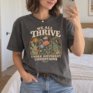 Könnte beinhalten: Ein anthrazitfarbenes Kurzarm-T-Shirt mit einem Grafikdruck aus drei gewölbten Feldern, die einen Kaktus, Blumen und Farne zeigen. Der Text lautet WE ALL THRIVE UNDER DIFFERENT CONDITIONS in einer cremefarbenen Schriftart.