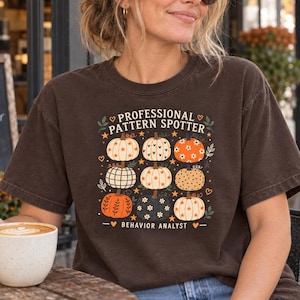 Op de afbeelding: Een donkerbruin T-shirt met korte mouwen en een afbeelding van negen pompoenen met diverse patronen. De tekst luidt PROFESSIONAL PATTERN SPOTTER en BEHAVIOR ANALYST, omringd door kleine sterren, blaadjes en hartjes in oranje en wit.