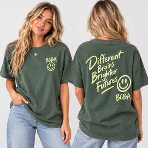 Könnte beinhalten: Ein dunkelgrünes Kurzarm-T-Shirt mit einem kleinen gelben Smiley und dem Text BCBA auf der Vorderseite. Die Rückseite zeigt den Text Different Brains Brighter Futures in gelber Schrift, begleitet von einem großen gelben Smiley und BCBA.