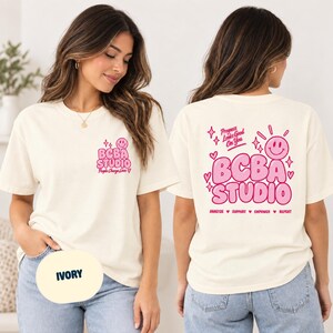 Könnte beinhalten: Ein elfenbeinfarbenes T-Shirt mit rosa Grafikdesigns. Die Vorderseite zeigt ein kleines Smiley-Gesicht und den Text BCBA STUDIO People Change Lives. Die Rückseite zeigt ein großes Smiley-Gesicht, den Text BCBA STUDIO, Progress Looks Good On You und ANALYZE SUPPORT EMPOWER REPEAT.
