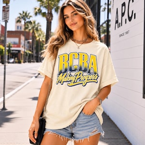 Könnte beinhalten: Ein cremefarbenes Kurzarm-T-Shirt mit dem Text BCBA in großen gelben und blauen Blockbuchstaben sowie dem Schriftzug Making Progress in blauer Schreibschrift darunter. Das T-Shirt hat einen Rundhalsausschnitt und eine lockere Passform.