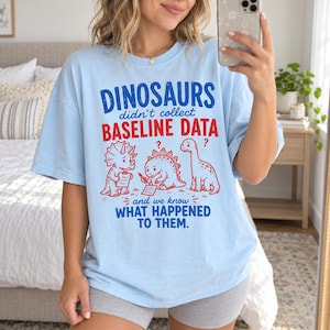 Op de afbeelding: Een lichtblauw t-shirt met rode en blauwe tekst die luidt DINOSAURS didn't collect BASELINE DATA en we know WHAT HAPPENED TO THEM. De afbeelding bevat drie lijntekeningen van dinosaurussen, waarvan er één een klembord vasthoudt.