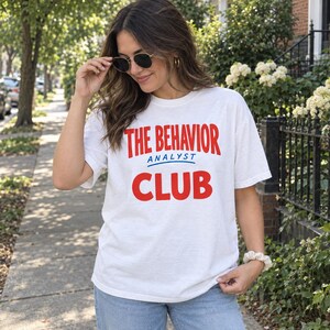 Könnte beinhalten: Ein weißes kurzärmeliges T-Shirt mit dem roten Schriftzug THE BEHAVIOR und CLUB sowie dem blau unterstrichenen blauen Wort ANALYST. Das T-Shirt hat einen Rundhalsausschnitt und eine lockere Passform, kombiniert mit einer hellblauen Jeans.