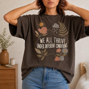 May include: A charcoal grey short-sleeve t-shirt featuring a graphic print with the text WE ALL THRIVE UNDER DIFFERENT CONDITIONS. The design includes illustrations of a sun, a rain cloud, ferns, and various flowers in muted earth tones.