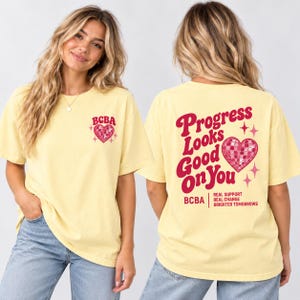Op de afbeelding: Een lichtgeel t-shirt met korte mouwen en een kleine roze discobal-hartafbeelding op de linkerborst met de tekst BCBA. De achterkant toont de tekst Progress Looks Good On You, een groot roze discobal-hart en de tekst BCBA.