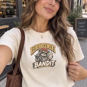 May include: A cream-coloured short-sleeve t-shirt featuring a graphic of a raccoon wearing a beanie and sunglasses, holding a coffee cup. The text above the raccoon reads REINFORCEMENT and the text below reads BANDIT in bold, distressed lettering.