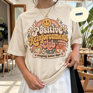 Op de afbeelding: Een ivoorkleurig t-shirt met korte mouwen en een grafische print met de tekst Positive Reinforcement Club en shaping better days. Het ontwerp bevat een geel smileygezicht, een regenboog, bloemen, een hart en een pot met een geruit deksel.