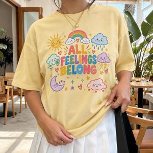 Könnte beinhalten: Ein hellgelbes Kurzarm-T-Shirt mit einem bunten Grafikdruck und dem Text ALL FEELINGS BELONG. Das Design zeigt eine lächelnde Sonne, einen Regenbogen, eine traurige Regenwolke, eine windige Wolke, einen violetten Mond, einen Blitz und Herzen.