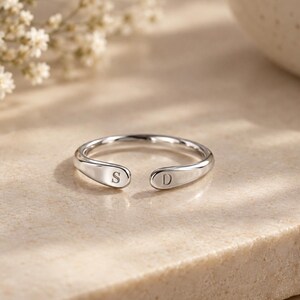 Könnte beinhalten: Ein silberfarbener offener Ring mit glatter, polierter Oberfläche, der an einem Ende mit dem Buchstaben S und am gegenüberliegenden Ende mit dem Buchstaben D graviert ist. Der Ring ist kreisförmig mit einer Lücke zwischen den Enden.