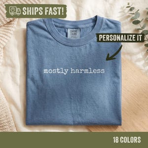 Peut inclure: Un t-shirt bleu à manches courtes avec une encolure ronde. Le vêtement présente le texte "mostly harmless" imprimé en blanc au centre de la poitrine. Une étiquette à l'intérieur du col indique "Comfort Colors". Le tissu a une texture mate.