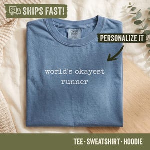May include: A blue short-sleeved t-shirt with a crew neckline. The center of the shirt features the white text "world's okayest runner" in a simple, clean font. The interior neck label displays the brand name Comfort Colors.