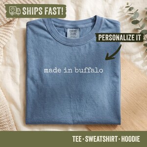 May include: A blue short-sleeved t-shirt with a crew neckline. The shirt features the white text "made in buffalo" printed across the chest. The interior neck label displays the brand name Comfort Colors in white text on a blue background.