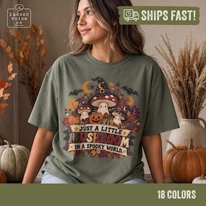 May include: An olive green t-shirt featuring a graphic of three smiling mushrooms wearing witch hats, surrounded by pumpkins, autumn leaves, and bats. The text reads, "JUST A LITTLE MUSHROOM IN A SPOOKY WORLD." The design includes stars and crescent moons.