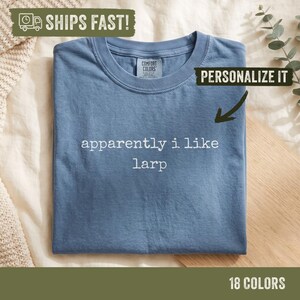 May include: A blue short-sleeved t-shirt with a crew neckline. The shirt features the white text "apparently i like larp" printed in the center. The inner collar tag displays the brand name Comfort Colors.