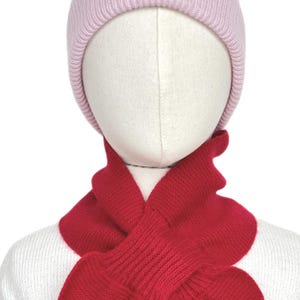 Puede incluir: Un gorro de punto rosa claro con un borde acanalado combinado con una bufanda de punto roja vibrante. La bufanda presenta un diseño de paso donde un extremo se inserta a través de una ranura en el otro, creando un aspecto superpuesto en el cuello.