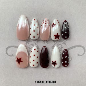 May include: A set of ten almond-shaped press-on nails featuring a variety of designs including white tips, red polka dots, solid dark maroon, and red star accents. The nails are arranged in two rows, with the text YUKARI ATELIER printed at the bottom.