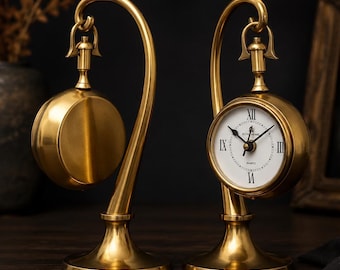 Könnte beinhalten: Zwei goldfarbene Tischuhren aus Metall, die an geschwungenen Ständern hängen. Eine Uhr zeigt ein weißes Zifferblatt mit schwarzen römischen Ziffern und Zeigern, die andere zeigt ihre schlichte, goldfarbene Rückseite. Beide haben ein rundes Design.
