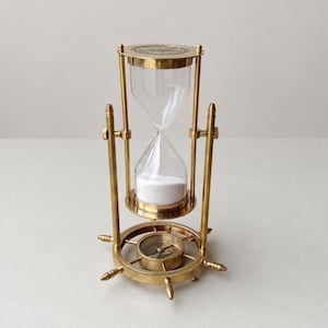 May include: A brass hourglass featuring a clear glass bulb filled with white sand, suspended within a rotating frame. The base is designed as a ship's wheel with a central compass, and the top cap features an engraved circular design.