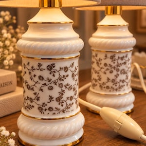 Może przedstawiać: Dwie lampy stołowe z białymi ceramicznymi podstawami z brązowym kwiatowym wzorem i złotymi wykończeniami. Podstawy mają skręcony wzór liny na górze i na dole. Każda lampa posiada szary abażur z tkaniny oraz biały przewód zasilający.