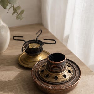 Pode incluir: Dois componentes de queimador de metal vintage para fogão portátil. A peça frontal é uma base circular em tom de cobre com um centro roscado e o texto "RECHAUD INUCCABLE A GAZ D'ALCOOL". A peça traseira é uma base em tom de latão com quatro pontas de metal preto.