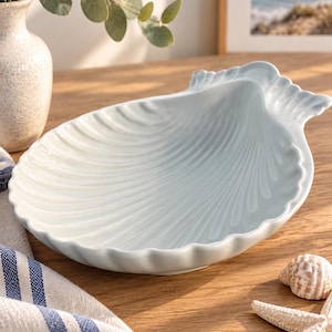 May include: A light blue ceramic dish shaped like a scallop shell with a glossy finish and raised ridges radiating from the center. The dish features a scalloped edge and a smooth, curved interior surface, designed as a decorative serving piece.