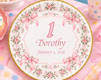 May include: A round paper plate with a white center and a gold rim. The design features a pink floral wreath with four pink bows. In the center, the number 1 is printed above the name Dorothy and the date January 1, 2026, in pink text.