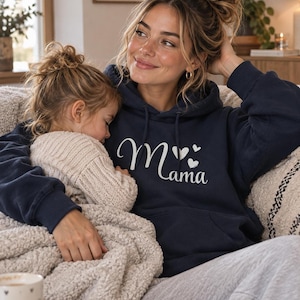 Könnte beinhalten: Ein marineblauer Kapuzenpullover mit dem weißen Schriftzug "Mama" auf der Brust, ergänzt durch drei kleine weiße Herzsymbole über dem Buchstaben "m". Das Kleidungsstück verfügt über eine Kängurutasche auf der Vorderseite und passende Kordelzüge.