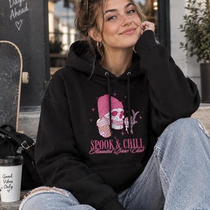 Könnte beinhalten: Ein schwarzer Kapuzenpullover mit einer Grafik eines Totenkopfes, der eine rosa Mütze trägt, einen Kaffeebecher hält und ein Friedenszeichen zeigt. Der Text lautet SPOOK & CHILL Haunted Brew Club in rosa Schrift. Der Hoodie hat lange Ärmel.